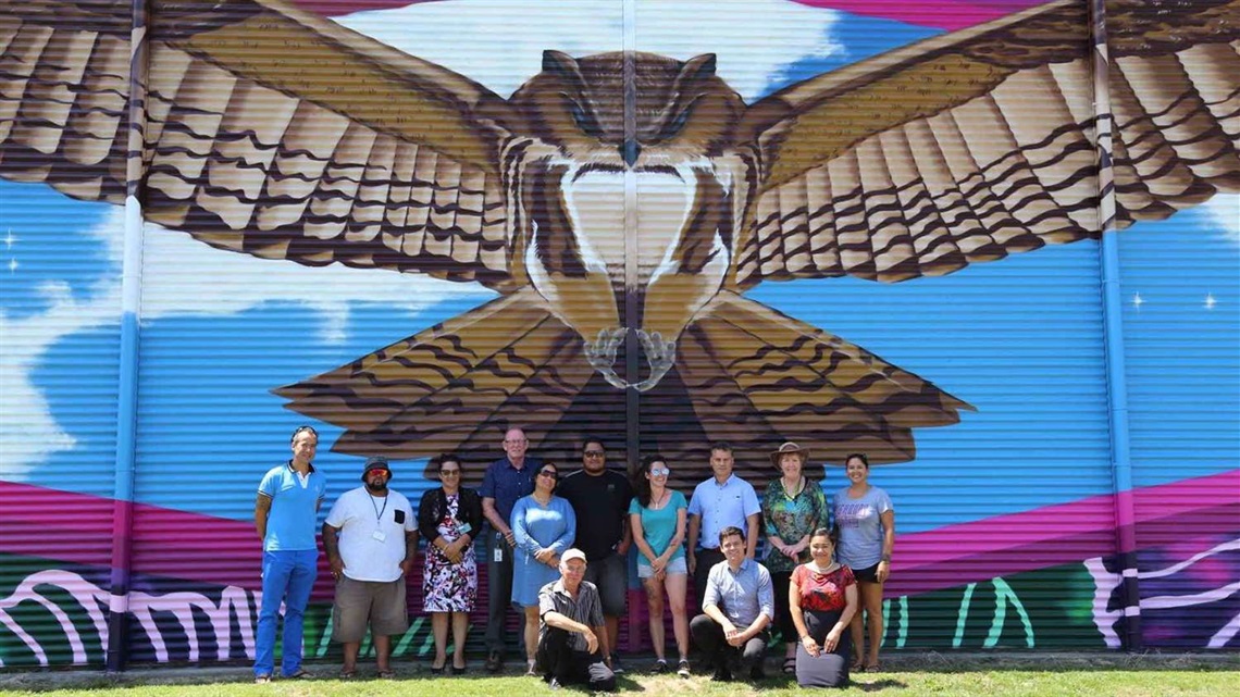 Smiling people grouped in front of a colourful mural depicting a native falcon with spread-eagled wings that appear to embrace them.