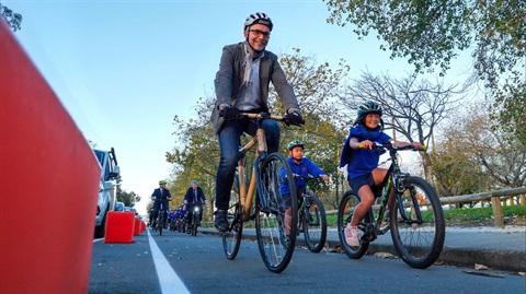 Smiling man and two primary school children biking along a cycleway separated from the road carriage by colourful planter boxes. 