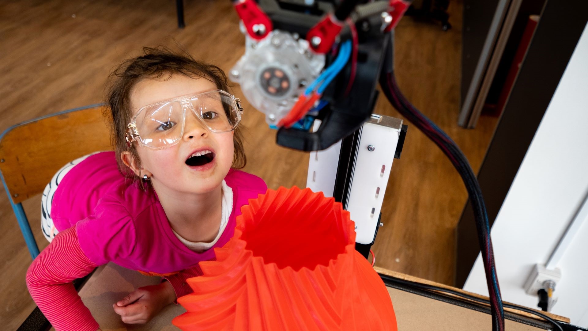 A small child wearing goggles is open-mouthed in wonder as the 3D printer forms a brightly coloured and sculptural vase.