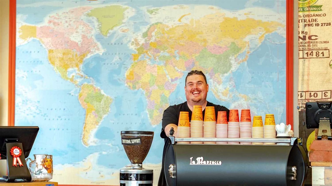 Smiling barista stands at espresso machine topped with compostable takeaway cups.