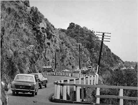 Black and white photo shows two cars travelling along the road by a gorge