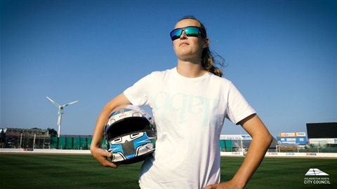 Photo shows young woman driver wearing reflective sunglasses on the speedway track holding her helmet.