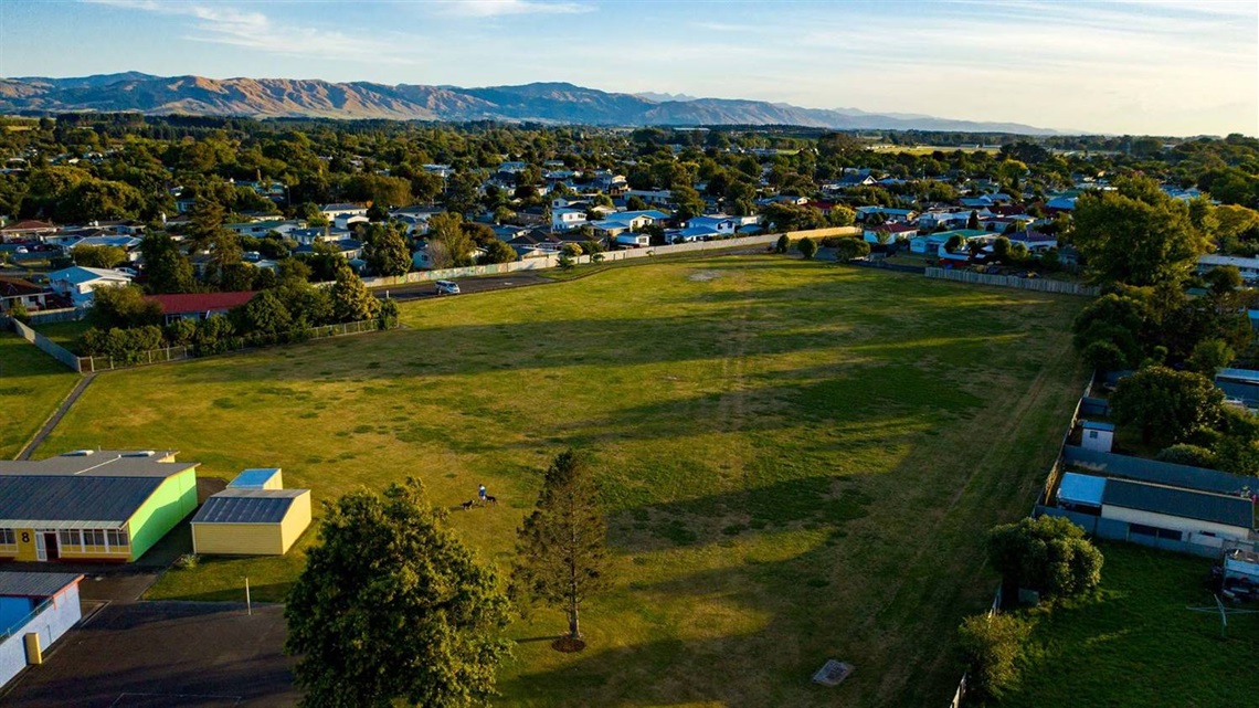 Photo shows a grassy field surrounded by houses. The hills are visible in the background.