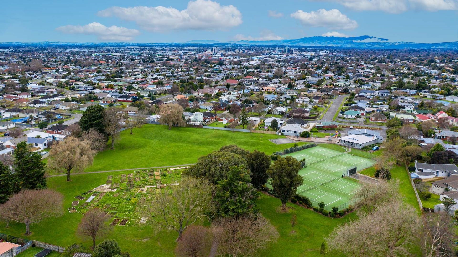 Photo shows aerial view of a grassy park with some established shade trees around the perimeter, a number of lawn tennis courts, and the community gardens. Neighbouring houses stretch into the background.