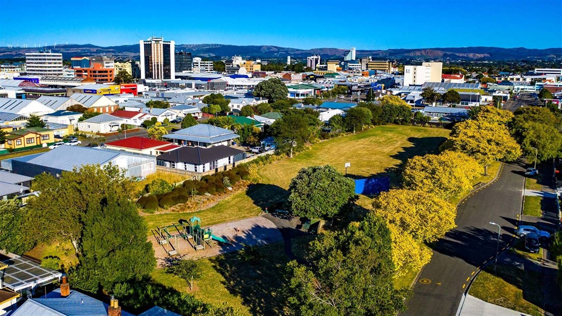 Photo shows small neighbourhood park with a few shade trees, some grass to run around, swings and a slide.