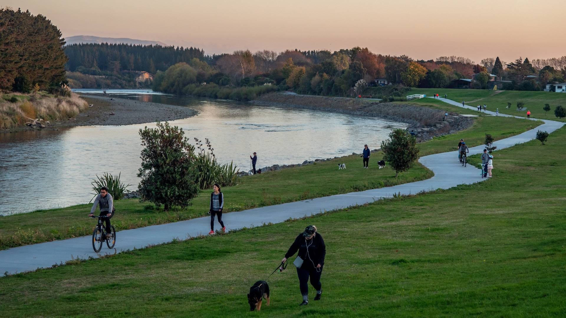 A busy walkway alongside the riverbank with lots of people biking, walking, and exercising dogs.