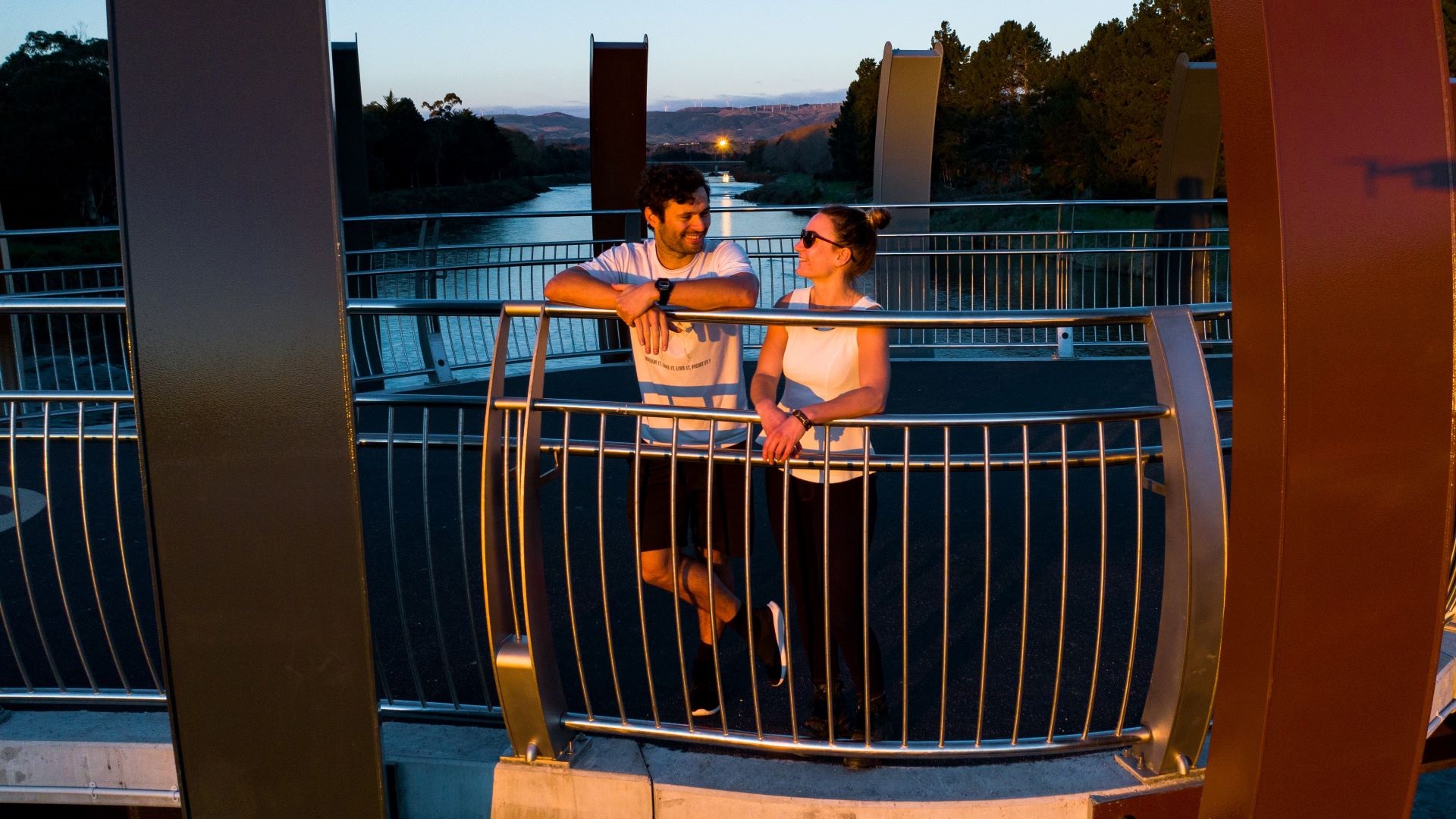 Photo shows young couple grinning at each other while they stand in the middle of the bridge at sunset with the river stretching out behind them.