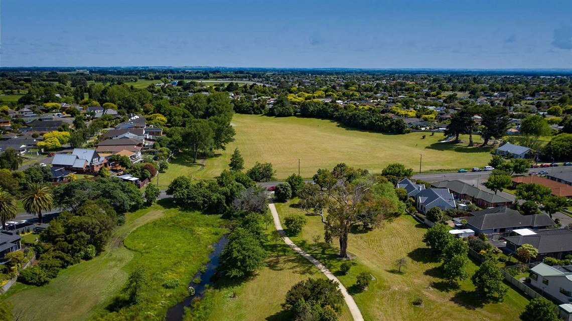 Photo shows aerial view of reserve with a stream and a limestone walkway running through it and a large flat grassed area that might be sportsfields in the background.