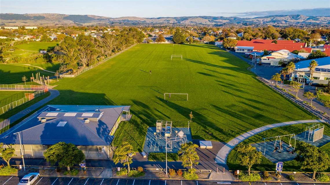 Photo shows aerial view of sportsfields and a small playground neighboured by a reserve, a large car park, and Freyberg swimming pool.