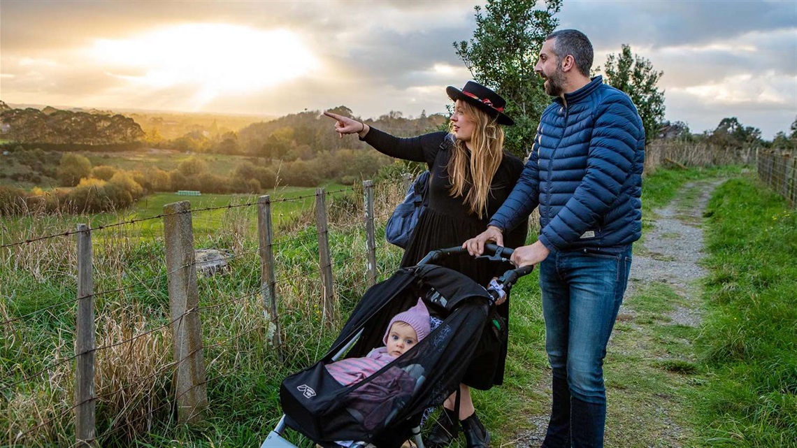 Photo shows young couple strolling with a baby in a pushchair, momentarily stopped to gaze out at the beautiful rural views. The sun is setting over trees in the distance.