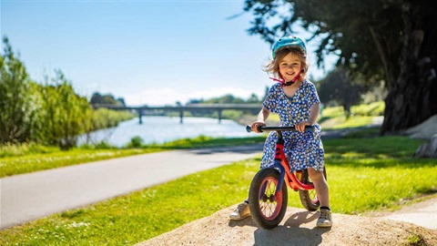 Smiling girl rides balance bike through the jumps alongside the river pathway.