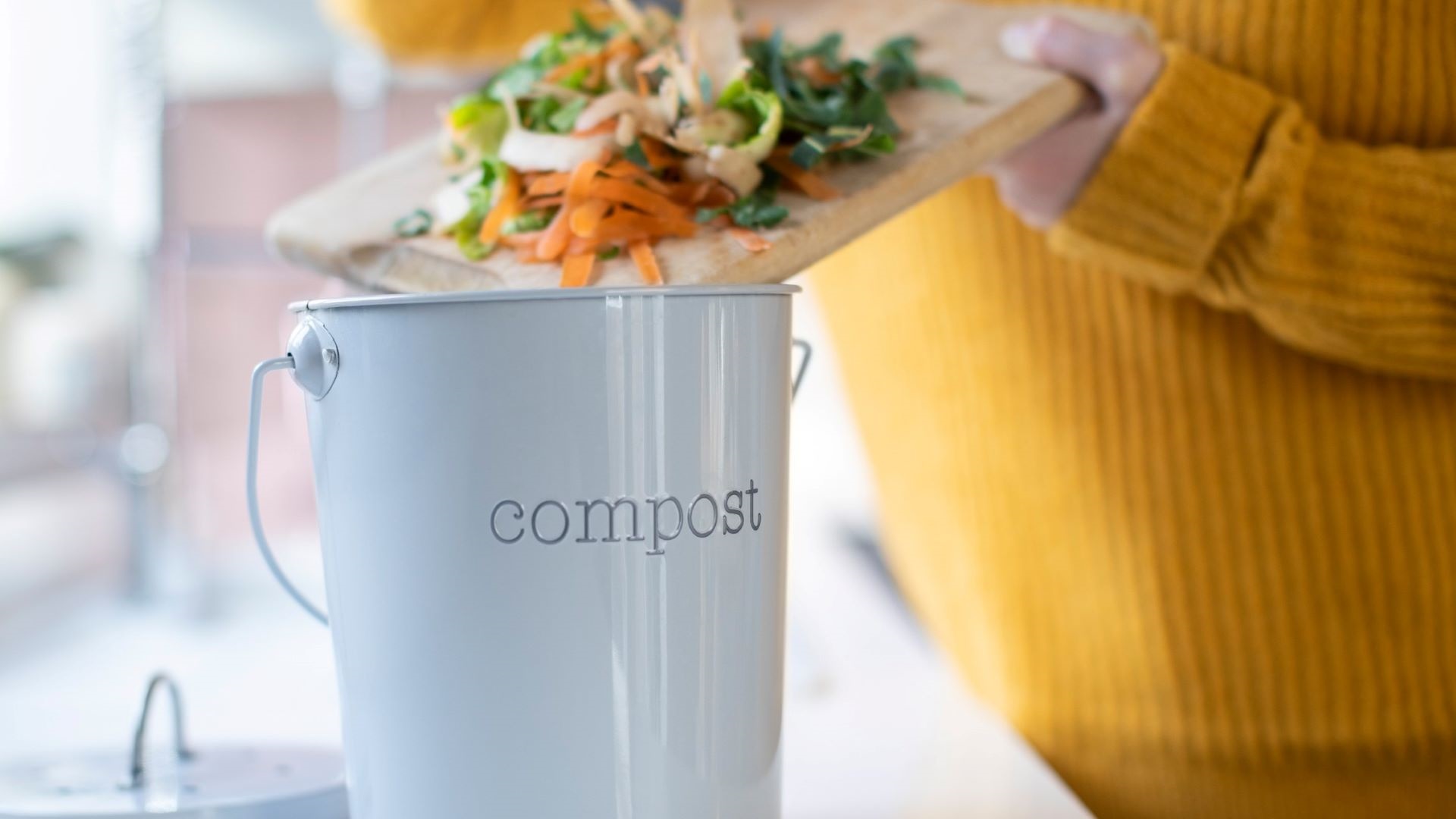 Photo shows person scraping vegetable peelings from a chopping board into a compost bin on the kitchen bench.