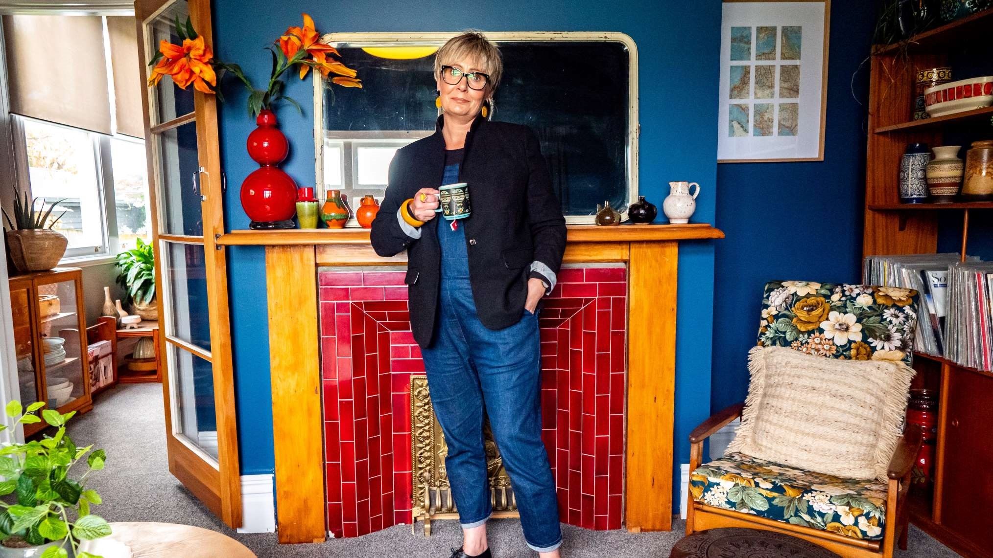 Photo shows stylish woman drinking from a Crown Lynn mug, standing in her colourful and comfortable home which is fully furnished with an eclectic array of second-hand treasures.