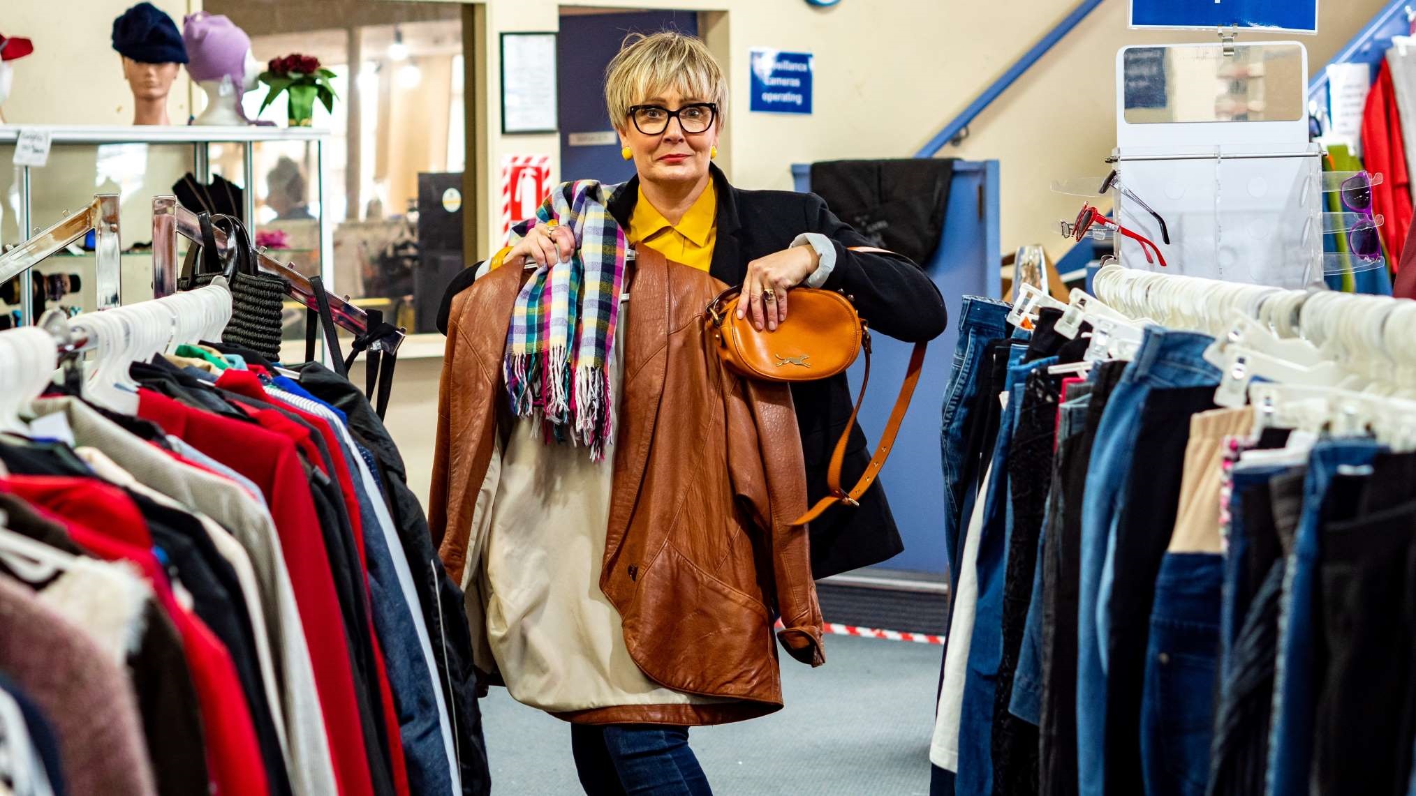 Photo shows stylish woman with bold spectacles standing in a clothing aisle at an op-shop, holding up an outfit she's pulled from the racks: leather jacket, scarf and bag.