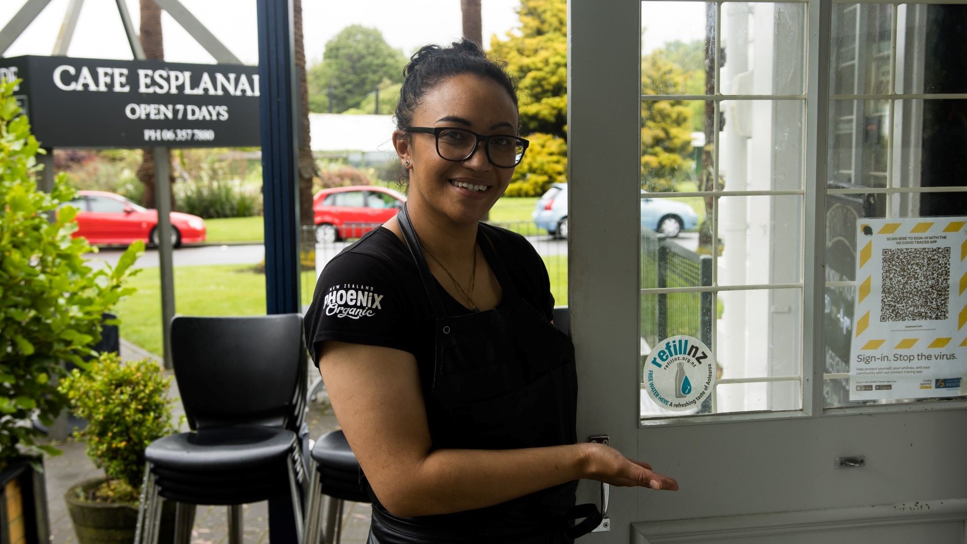 Photo shows smiling woman showing off Refill NZ sticker displayed in the window of her cafe.