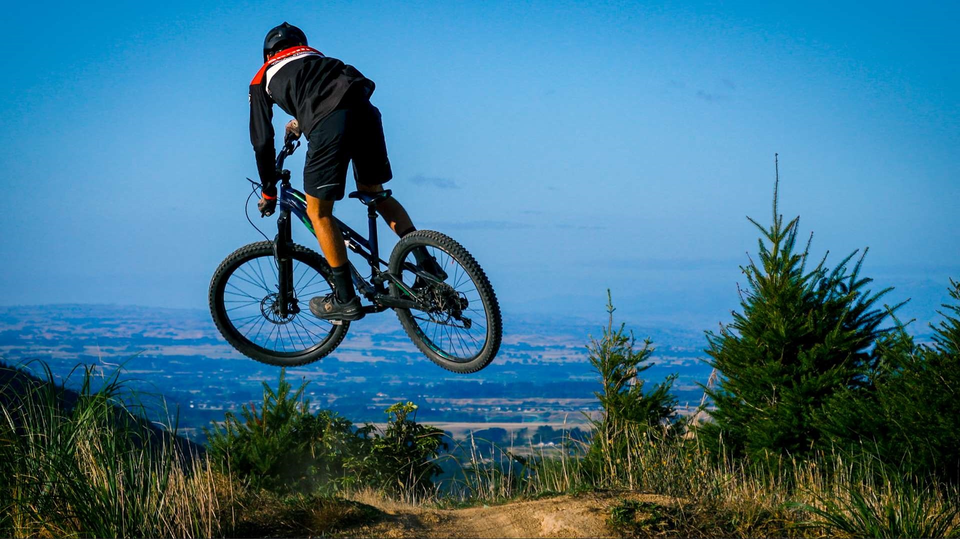 Photo shows man on mountainbike leaping in the air on a dirt track at the top of a hill, with views out towards Palmerston North.