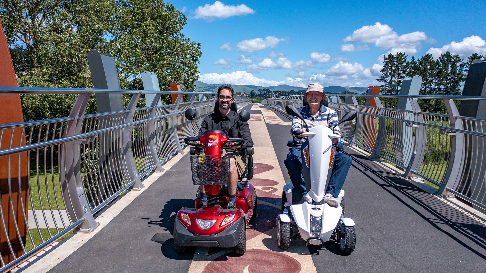 Two men are riding mobility scooters on the He Ara Kotahi bridge.