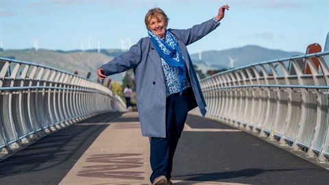 Older woman smiling with her arms outstretched while walking across a pedestrian bridge.