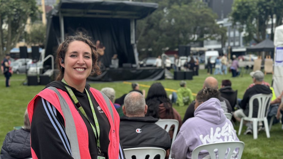 Woman in a high-vis vest smiles at the camera at an outdoor event, with a small stage and seated audience on a grassy lawn behind her.