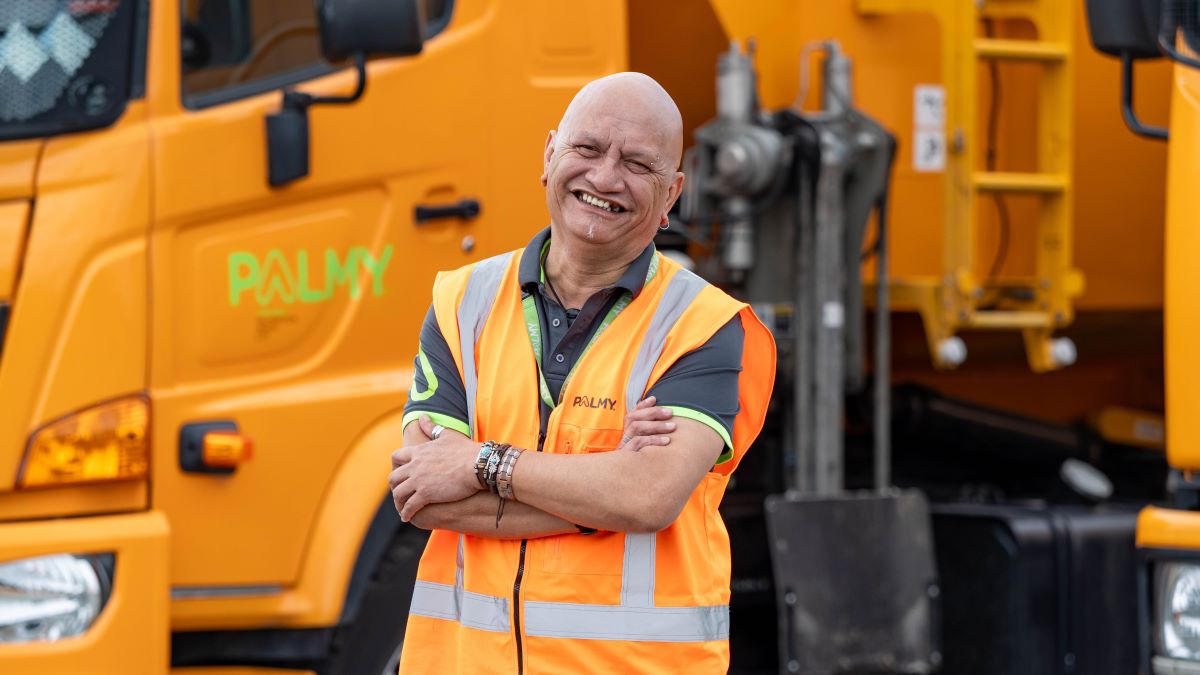 A smiling man standing with folded arms in front of a rubbish collection truck.