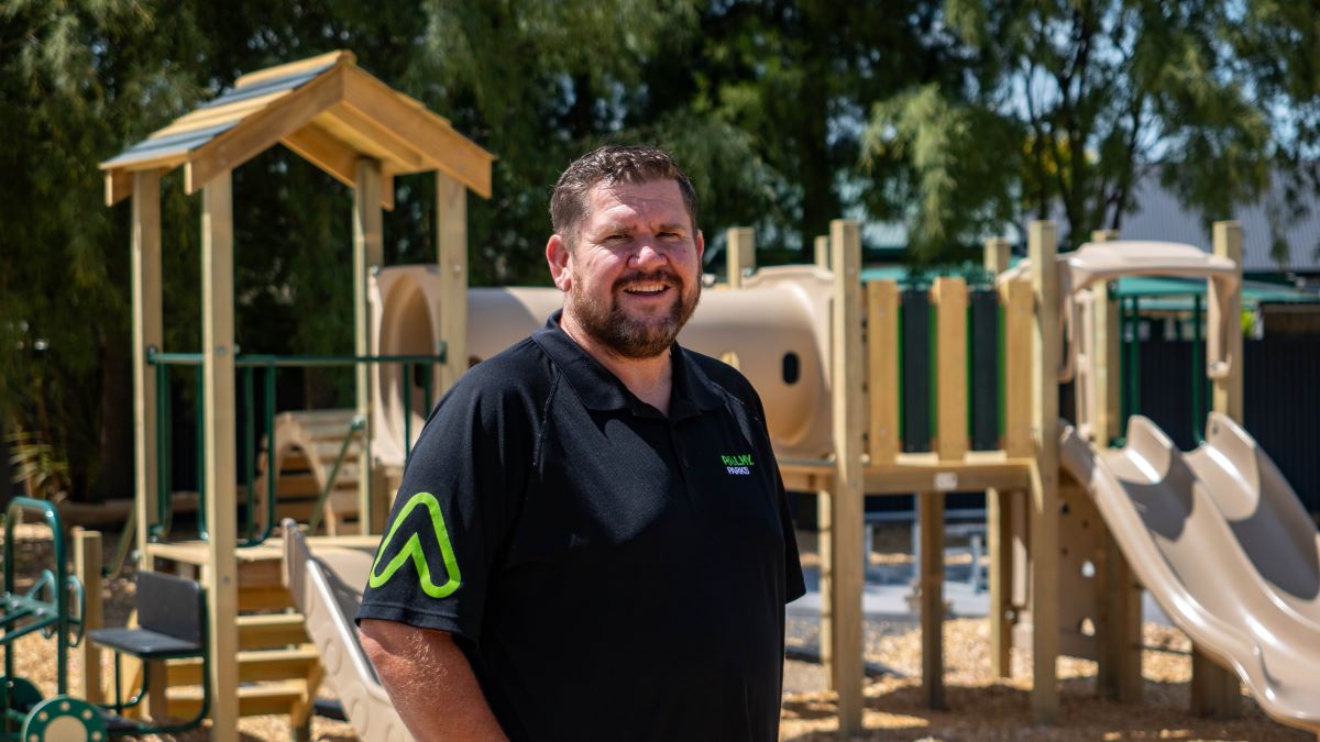 A smiling, bearded man in a children's playground with a cool structure in the background for climbing, sliding, spinning and sparking imaginative play.