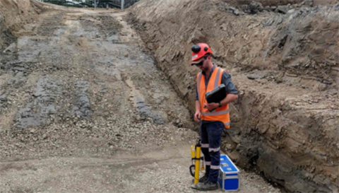 A worker in safety gear standing at the bottom of a large hole, clegg hammer testing.