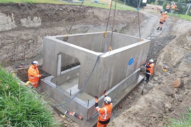 Workers inside the hole stand at each corner of a large concrete structure being carefully lowered in by crane.