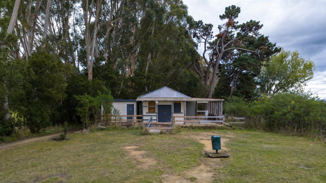 Small, weathered building with a tin roof and wooden deck, set in a grassy clearing surrounded by tall trees.