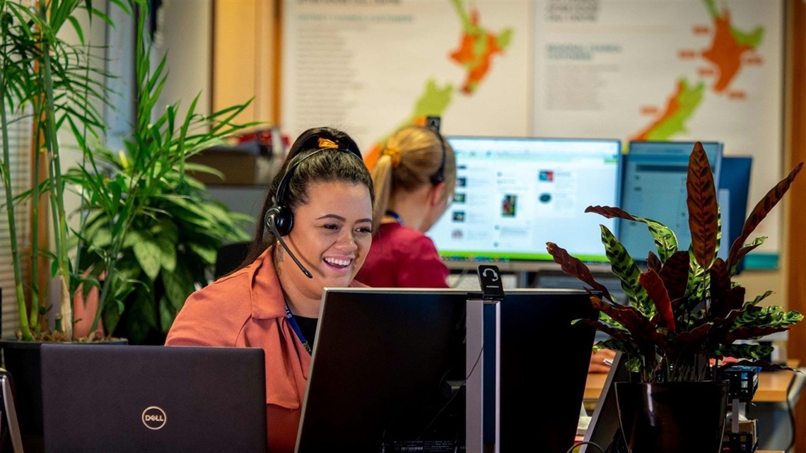 A smiling young woman wearing a headset logs a request for service on the computer in front of her while she takes a call from a resident.