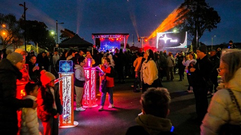Image shows people gathering around the colourful light poles, with screens and stages at background