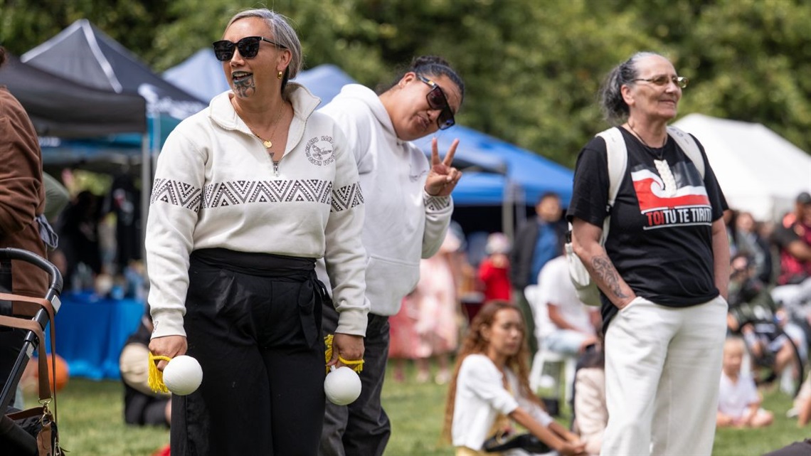 People enjoying Waitangi Day celebrations, with one person holding poi and stalls in the background.