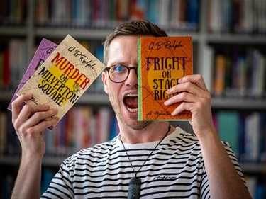image shows a man holding three novels in front of a bookshelf