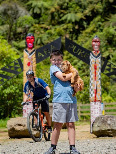image shows a boy holding his dog on a gravel cycling pathway, with a man riding on a mountain bike behind