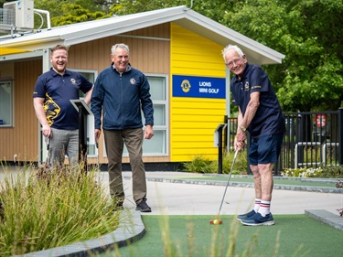 image shows three men playing minigolf in front of a yellow club house