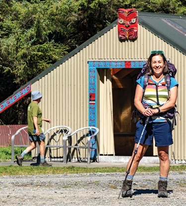 image shows a happy woman with hiking gears standing in front of a house
