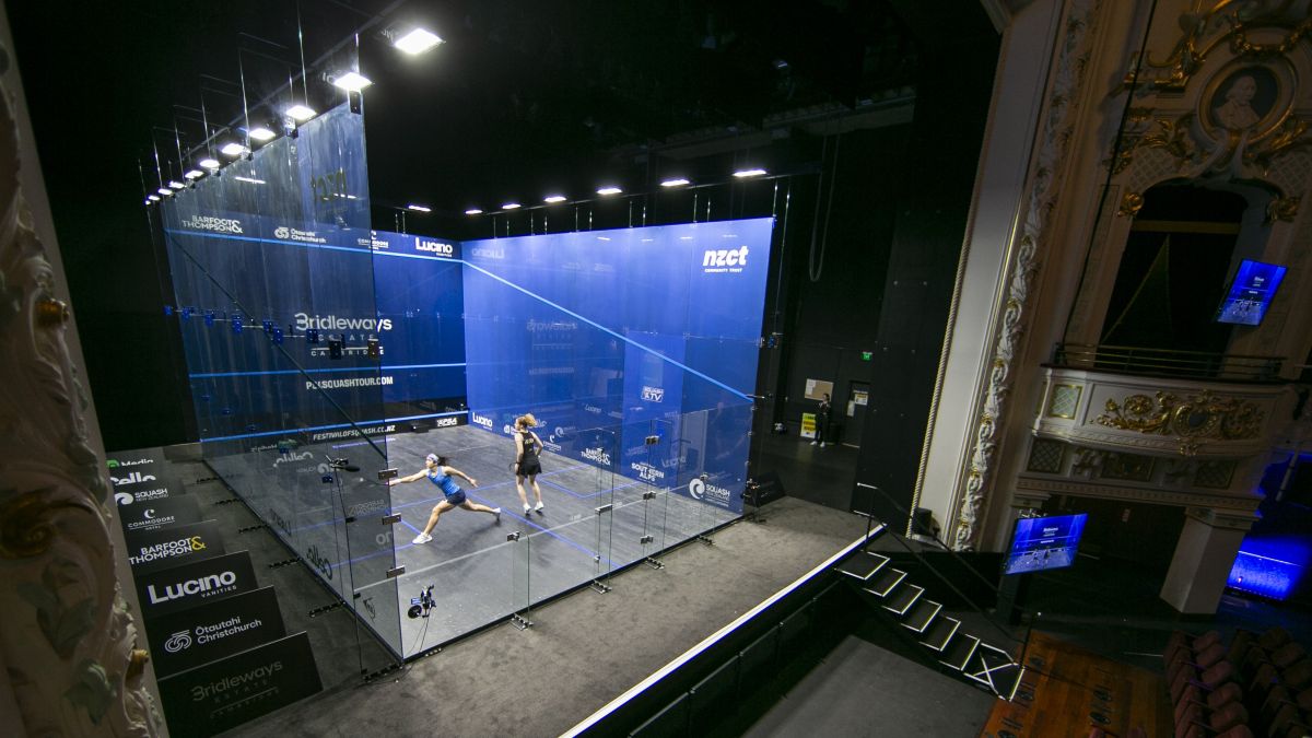 Two women play squash in a glass-walled court on a raised theatre stage.