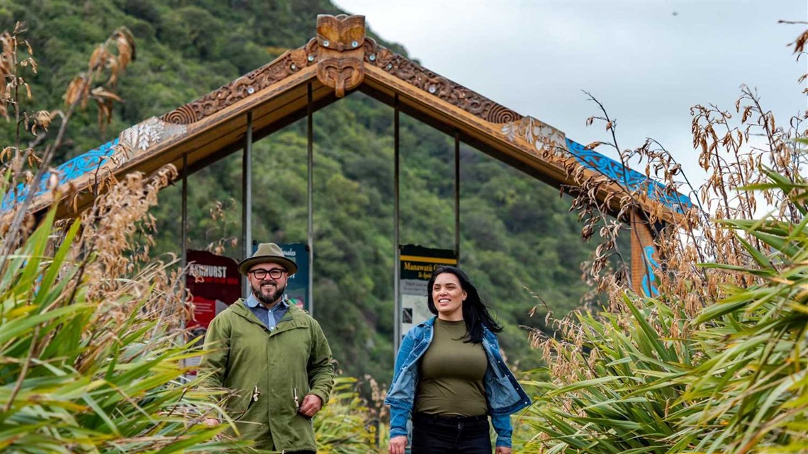 A smiling man and woman walk along a flax-lined pathway with a carved structure in the background.