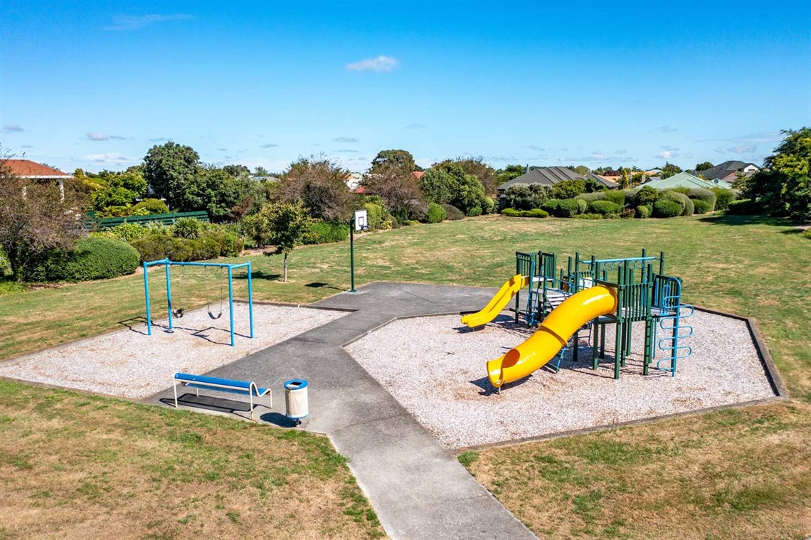 Image shows a playground and a basketball hoop at the centre of grass, surrounded by a bush.