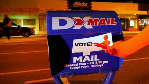 Photos shows an orange hand holding voting paper by a DX Mail box