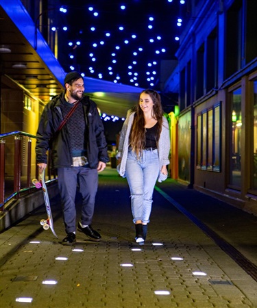A young couple stroll through a laneway at night festooned with lights.