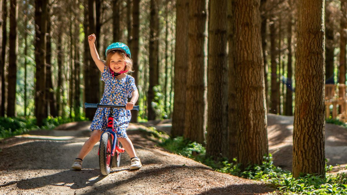 A smiling girl pumps her first in the air as she negotiates her balance bike over a bumpy track through the trees.