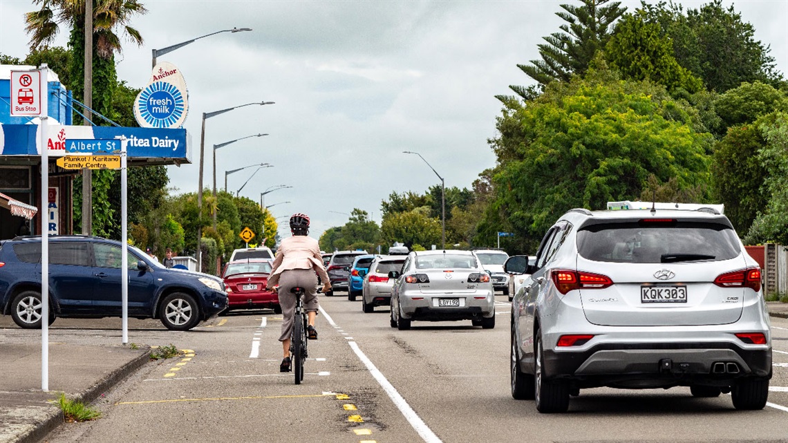 Woman biking towards a cross, dwarfed by passing vehicles on the painted bike lane.