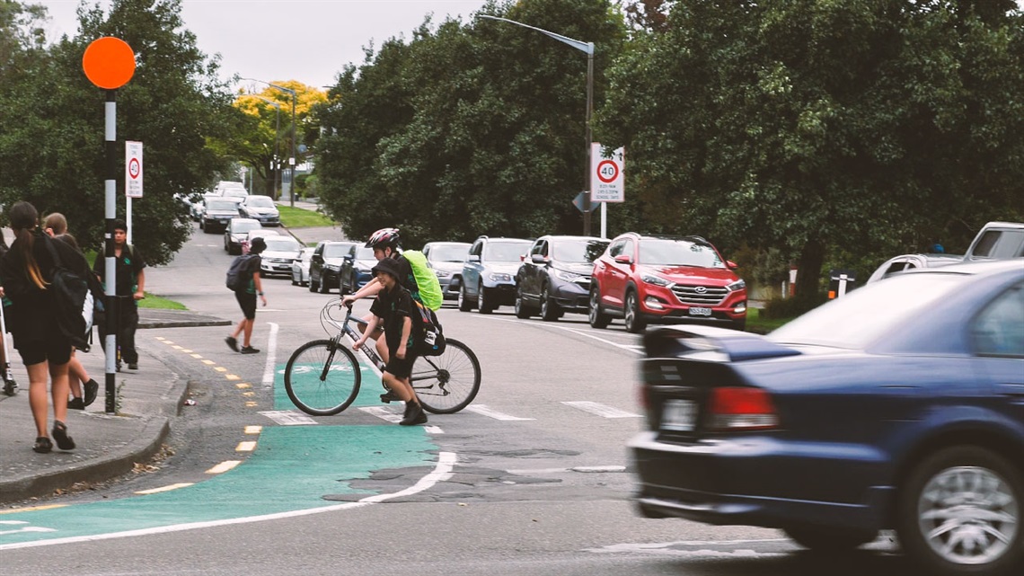 Image shows students walking or cycling across a busy road with cars parking on roadside