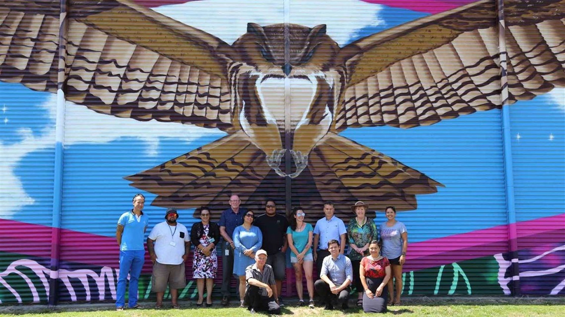 Smiling people grouped in front of a colourful mural depicting a native falcon with spread-eagled wings that appear to embrace them.