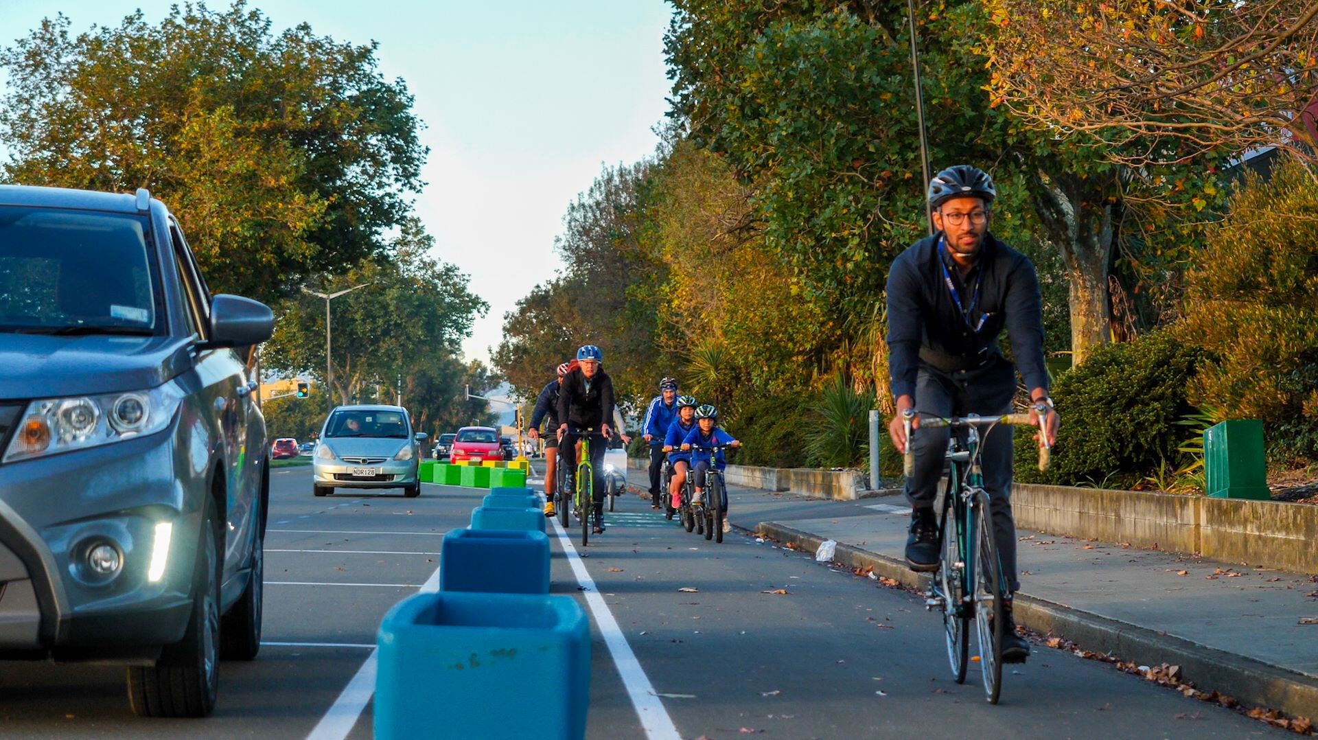 Group of adults and primary school kids biking along a cycleway that uses planter boxes to separate them from the cars.