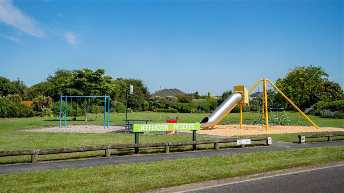 Image shows a playground and a basketball hoop at the centre of grass, surrounded by a bush.