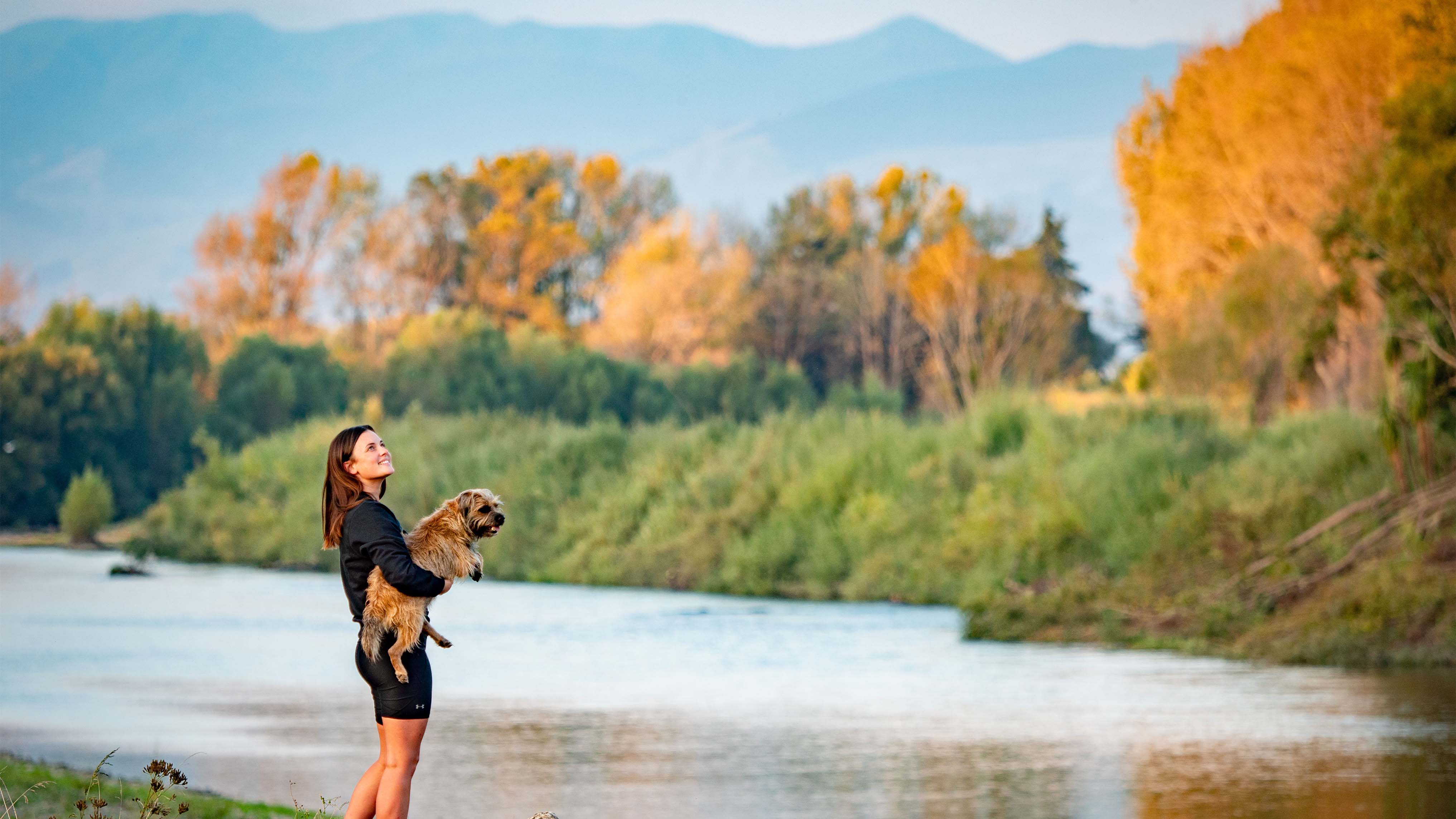 A woman standing on the pathway next to the riverbank, holding a small shaggy dog and gazing out over the water to the trees on the opposite bank.