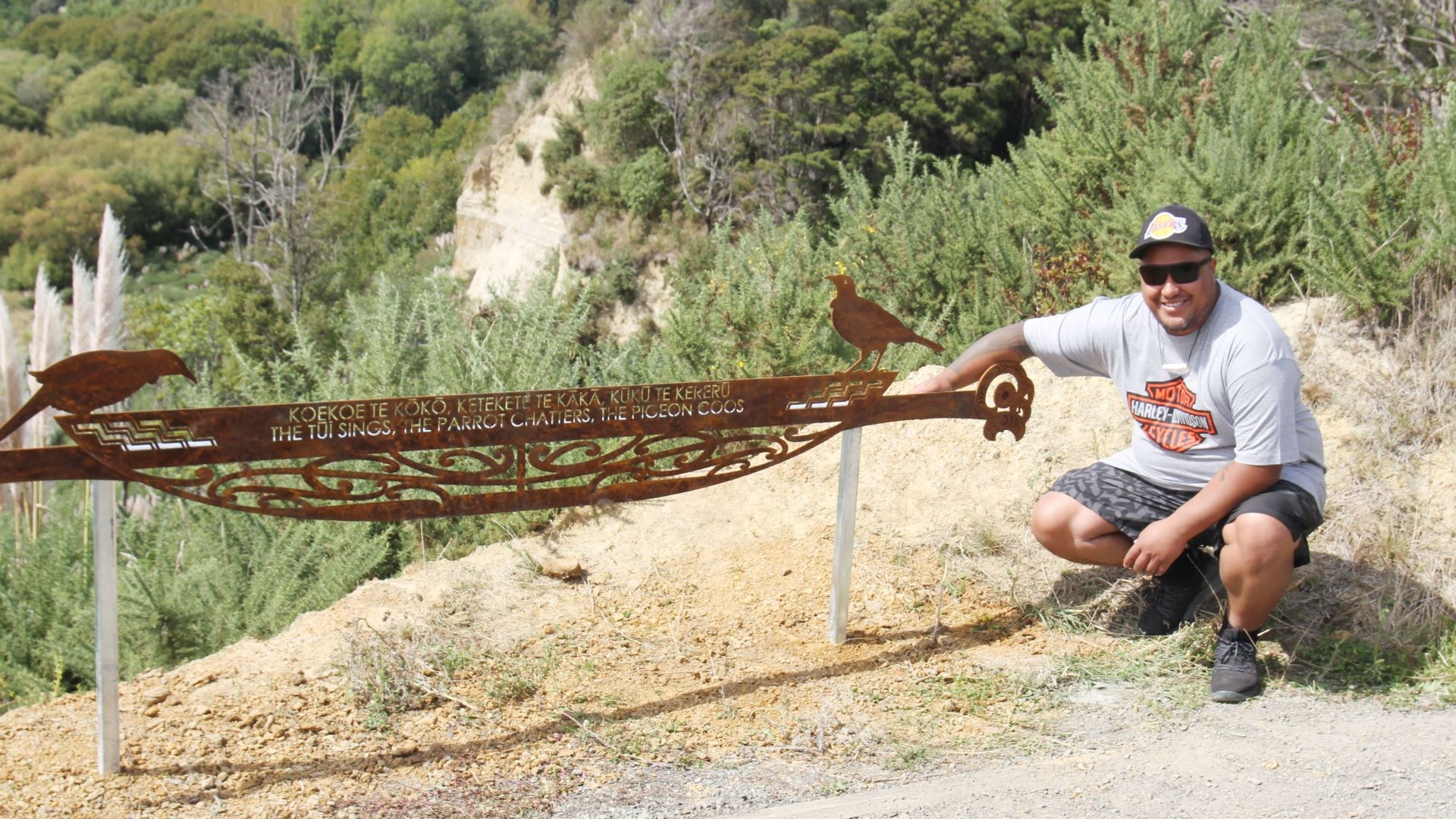 Photo shows the artist with a metalwork sign depicting native birds and one of the proverbs.
