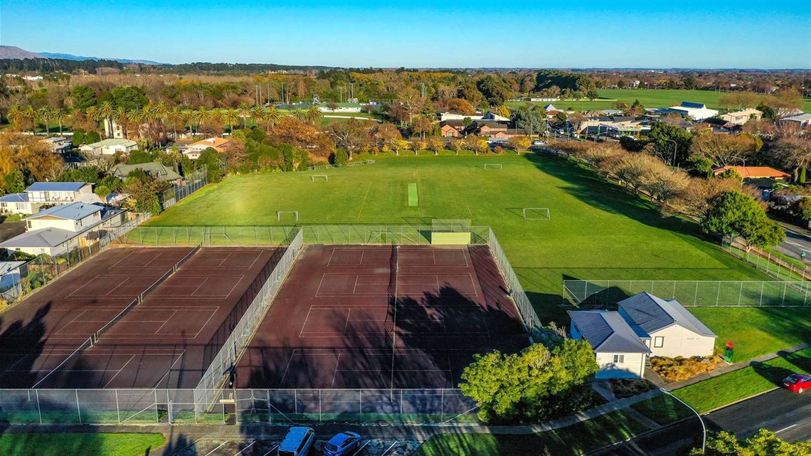 Aerial view of sportsfields lined with shade trees along one side and hard surface tennis courts in the foreground.