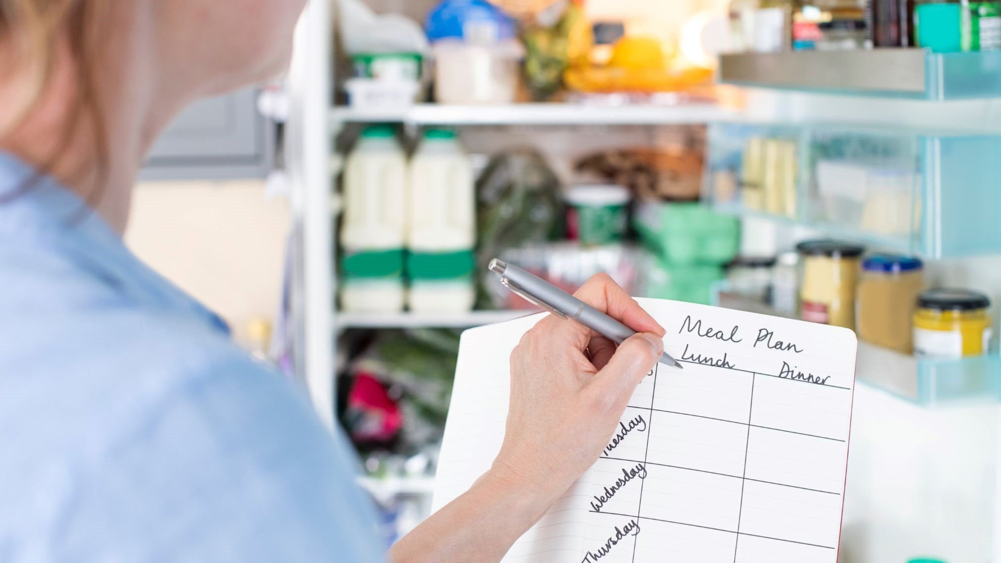 Woman standing in front of an open fridge with a notebook, writing her meal plan for the week.
